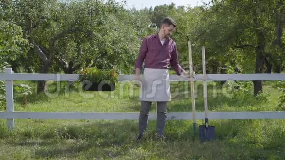 职业年轻的农民戴着花园手套站在绿色的夏季花园靠近栅栏那个人在视频的预览图