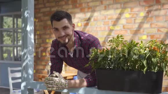 一幅年轻的微笑着的胡子农夫在门廊附近的一盆花上的画像看着摄像机微笑地站在里面视频的预览图