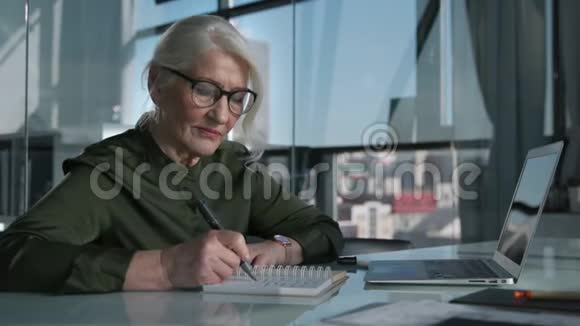 老妇人在办公室工作在笔记本上写字使用笔记本电脑视频的预览图