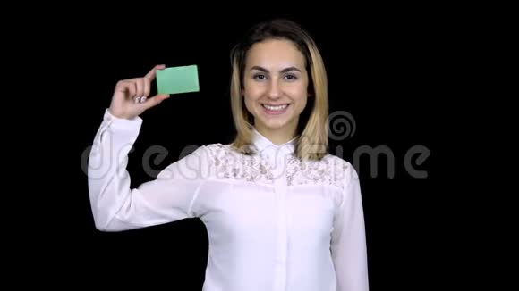 穿着白色衬衫的年轻女商人手里拿着一张银行卡孤立的黑色背景彩色绿卡4k视频的预览图