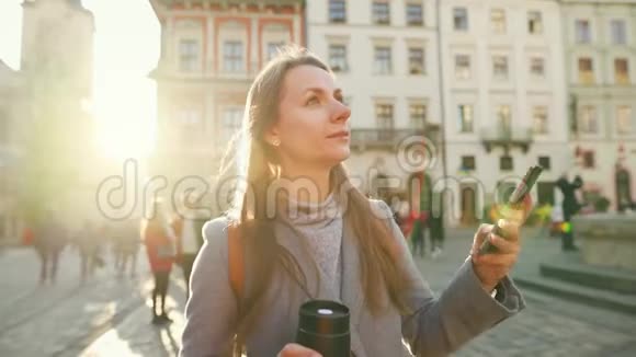 年轻的微笑女子穿着灰色外套手里拿着一个保温杯在漂亮的建筑智能手机上拍照视频的预览图