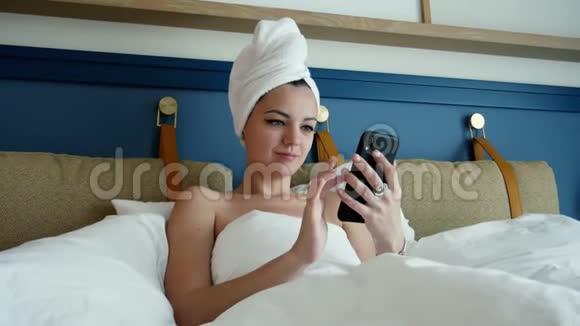 淋浴后躺在床上的漂亮性感年轻女子头上戴着毛巾使用智能手机滚动页面和视频的预览图
