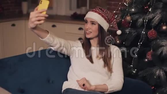 戴着圣诞帽坐在沙发上做自拍或做自拍的快乐白种人少女视频的预览图