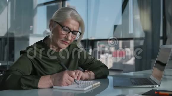 老妇人在办公室工作在笔记本上写字使用笔记本电脑视频的预览图
