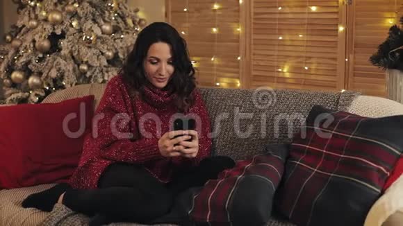 美丽的年轻女人用她的智能手机打字聊天笑她坐在灰沙发的圣诞树旁视频的预览图