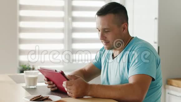年轻人在数字平板电脑上阅读新闻坐在厨房的窗户旁视频的预览图