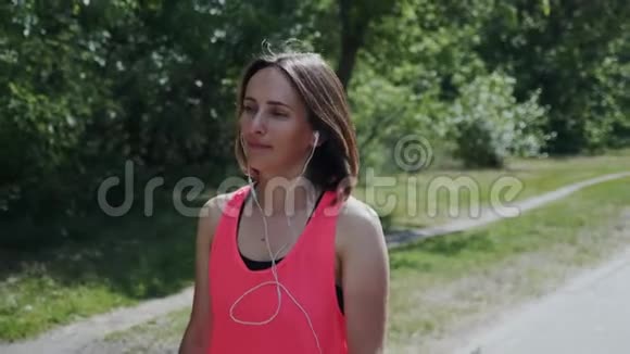 戴着耳机在公园散步的年轻快乐女孩有魅力的女人喜欢散步女孩跑步后放松布里的漂亮姑娘视频的预览图