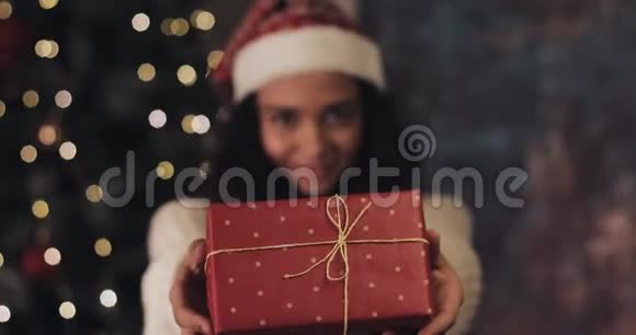 圣诞老人帽子里的快乐美丽的穆拉托女孩献礼和带回礼品盒看着镜头和微笑站在视频的预览图