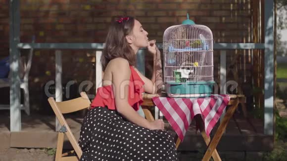 漂亮的女人穿着复古的连衣裙坐在桌子旁有一个笼子里面有两只鹦鹉在户外和鸟儿说话老式风格视频的预览图
