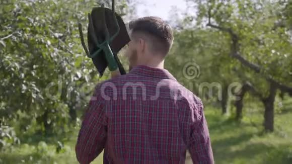 年轻农民肩上扛着一把铲子和一把草叉走过花园的后景农村生活观念视频的预览图