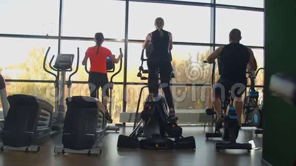 从后面看在健身房的有氧运动区妇女坐着椭圆机男子坐着运动自行车列车视频的预览图