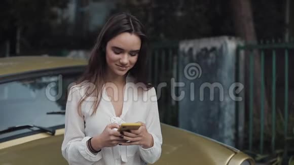 年轻迷人的布鲁内特高加索女孩站在老时尚的黄金车同时与朋友聊天通过视频的预览图