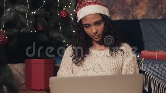 美丽的年轻女孩戴着桑塔帽坐在圣诞树下使用她的笔记本电脑打视频电话的女孩视频的预览图
