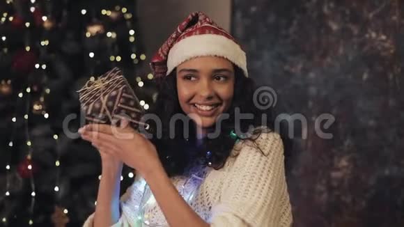 年轻漂亮的女士戴着桑塔的帽子脖子上挂着装饰灯手里拿着礼品盒看着镜头视频的预览图