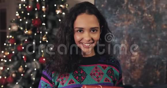 迷人的女人的肖像带着灿烂的微笑站在圣诞树旁拿着礼物盒微笑着看着视频的预览图