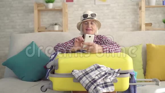 一个戴帽子的老女人微笑着手里拿着旅行箱的智能手机坐在沙发上视频的预览图