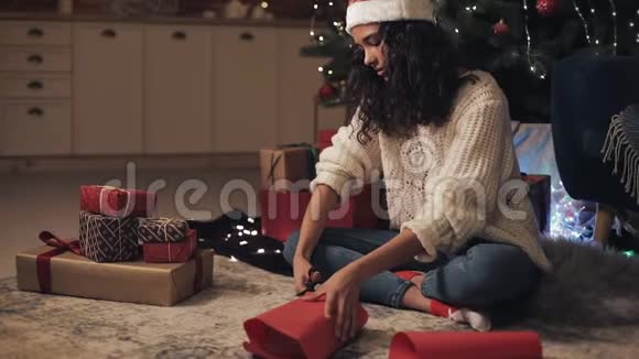 观看迷人的快乐穆拉托女孩在桑塔斯帽子准备礼物她剪纸包装礼物坐着视频的预览图