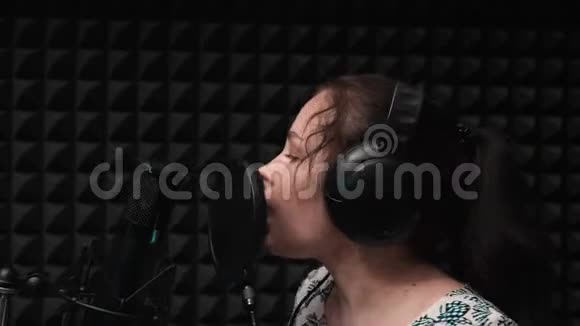 漂亮的女孩戴着蓝眼睛的耳机在录音室的麦克风附近唱歌录音激情歌唱视频的预览图