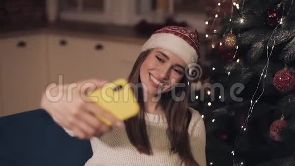 一个年轻迷人的女孩微笑着戴着桑塔帽坐在圣诞树附近的沙发上摆着自拍吹着一个吹着圣诞老人的帽子视频的预览图