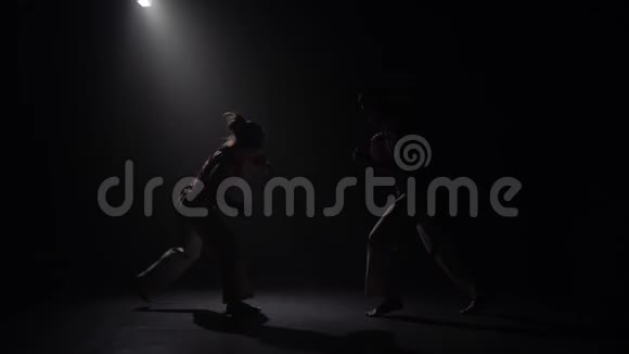一对夫妇在黑暗中练习卡波伊拉在演播室的聚光灯下慢动作视频的预览图