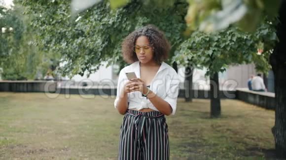 在城市公园户外使用智能手机的快乐非裔美国女孩画像视频的预览图