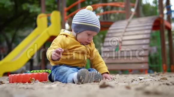 小可爱的男孩是坐在沙滩背景上玩耍的操场穿着黄色毛衣和帽子的秋天视频的预览图