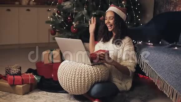 戴着桑塔斯帽子的年轻漂亮女人在笔记本电脑上打视频电话她坐在圣诞树附近的地板上视频的预览图