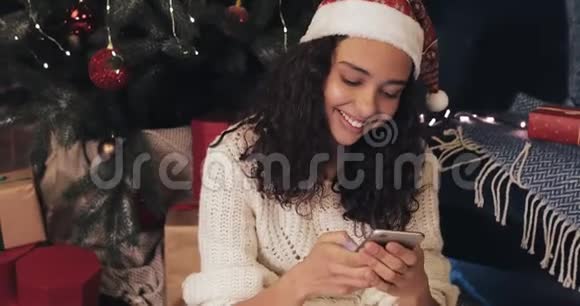 一个笑容可掬的年轻女孩在圣诞树附近的现代手机上发短信在社交网络上聊天视频的预览图