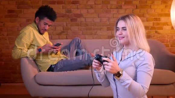 非洲男人在沙发上用智能手机工作白种人金发女孩在家里用操纵杆在地板上玩电子游戏视频的预览图