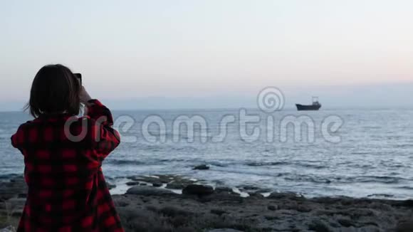 漂亮的黑发女孩在拍旧船的照片年轻的女孩穿着红色格子衬衫在黄昏时拍摄大海的照片女人拍照视频的预览图