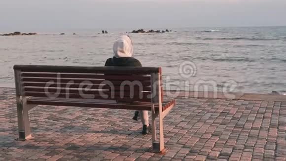 穿着黑色夹克衫头罩坐在海滩附近的长凳上的年轻女人女人看着暴风雨的大海坐在长凳上下雨的女孩视频的预览图