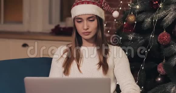 犹豫不决的年轻貌美的女孩坐在圣诞树旁用她的笔记本电脑思考看着视频的预览图