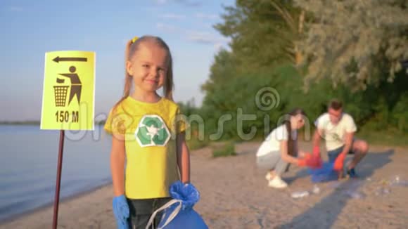 清洁海岸戴橡胶手套的小快乐志愿者女孩的肖像背景上的指针标志附近有垃圾袋视频的预览图