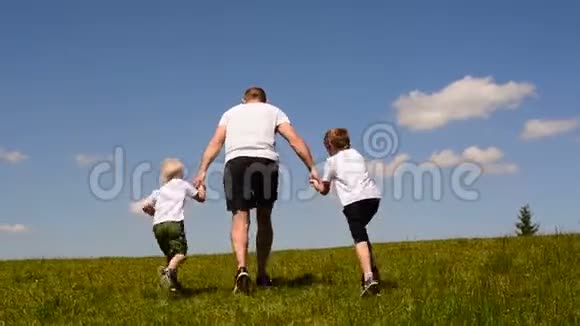 父亲和两个小儿子牵着手在绿色的草坪上奔跑天边乌云密布的蓝天视频的预览图