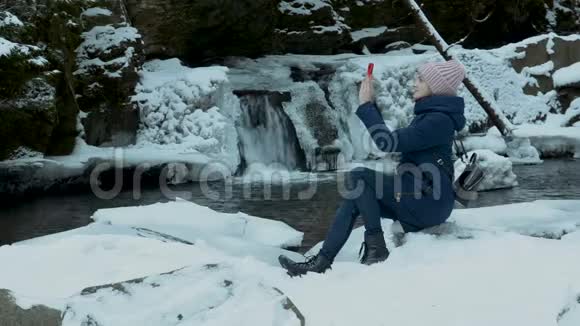 年轻女子坐在一个小瀑布的岸边在智能手机上拍摄视频冬季视频的预览图
