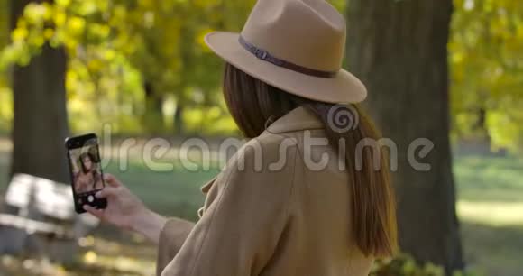年轻的高加索女孩穿着棕色帽子和外套在智能手机上自拍令人愉悦的欧洲女性消费视频的预览图