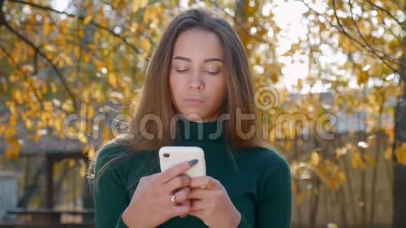 秋天年轻漂亮的黑发女人在黄树的背景下使用白色智能手机视频的预览图