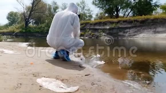 穿着防护服和呼吸器的人蹲在污染的河流湖泊旁用试管取水进行分析视频的预览图