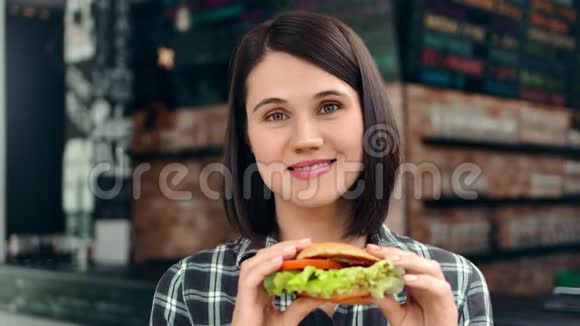 美丽的脸庞友好的年轻女孩摆姿势拿着新鲜多汁的汉堡看着镜头视频的预览图