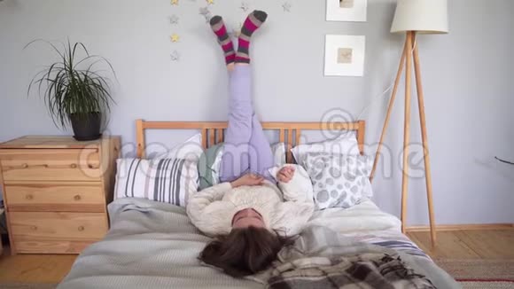 一个快乐的年轻女人躺在床上双腿靠在墙上到处乱跑穿着家庭服装休息五颜六色视频的预览图