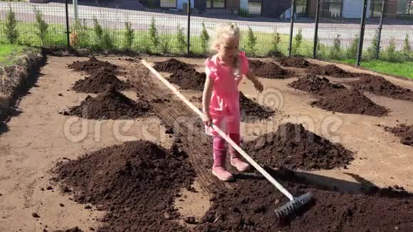 可爱的女孩穿着粉红色的衣服用耙子铲平了院子里的泥土视频的预览图
