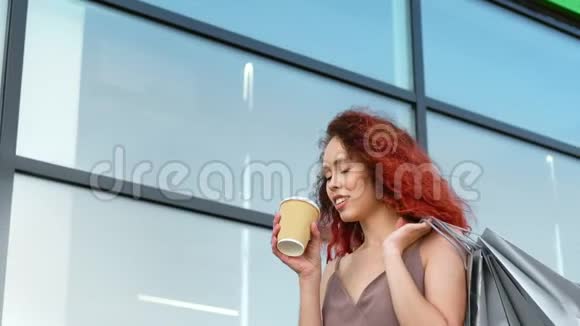 时尚购物狂红发女孩喝咖啡纸杯走窗玻璃视频的预览图