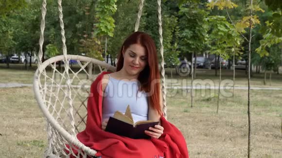 一个红头发的年轻女孩正在读一本书坐在一个城市公园里的一个白色悬挂的秋千上她微笑着视频的预览图