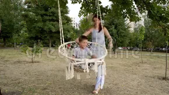 一个快乐的妈妈在城市公园的白色秋千上摇动她的小儿子他们在晴朗的秋日笑着玩这就是视频的预览图
