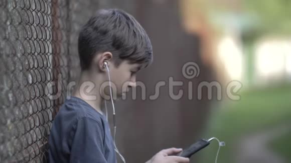 戴着耳机的有趣男孩站在一个带电话的围栏附近环顾四周在户外玩游戏视频的预览图