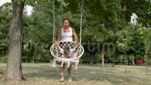 一位快乐的年轻母亲在一个明亮的夏天在公园里的白色秋千上摇动她的小儿子他们笑着玩视频的预览图