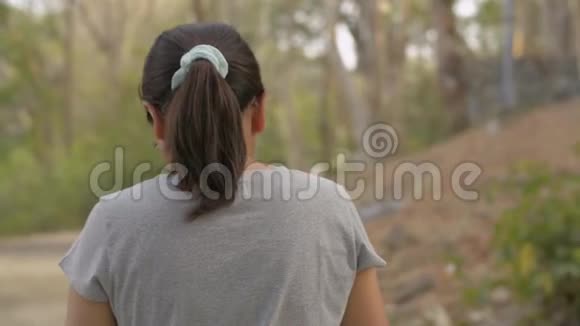 从后面看亚洲女人戴眼镜穿着休闲装使用手机悠闲地在自然公园散步视频的预览图