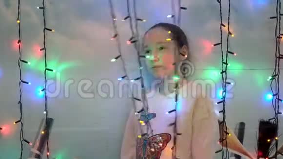 一个孩子一个小女孩站在房子墙附近的彩色花环后面她摸着她的手玩着视频的预览图