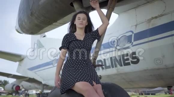 自信的年轻黑发女孩穿着黑色连衣裙看着坐在白色飞机起落架上的摄像机视频的预览图