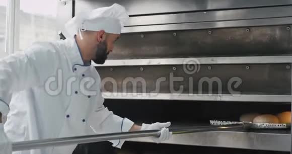 有魅力的面包师带着胡须用特殊的火炬从工业烘炉机上取出煮熟的面包视频的预览图